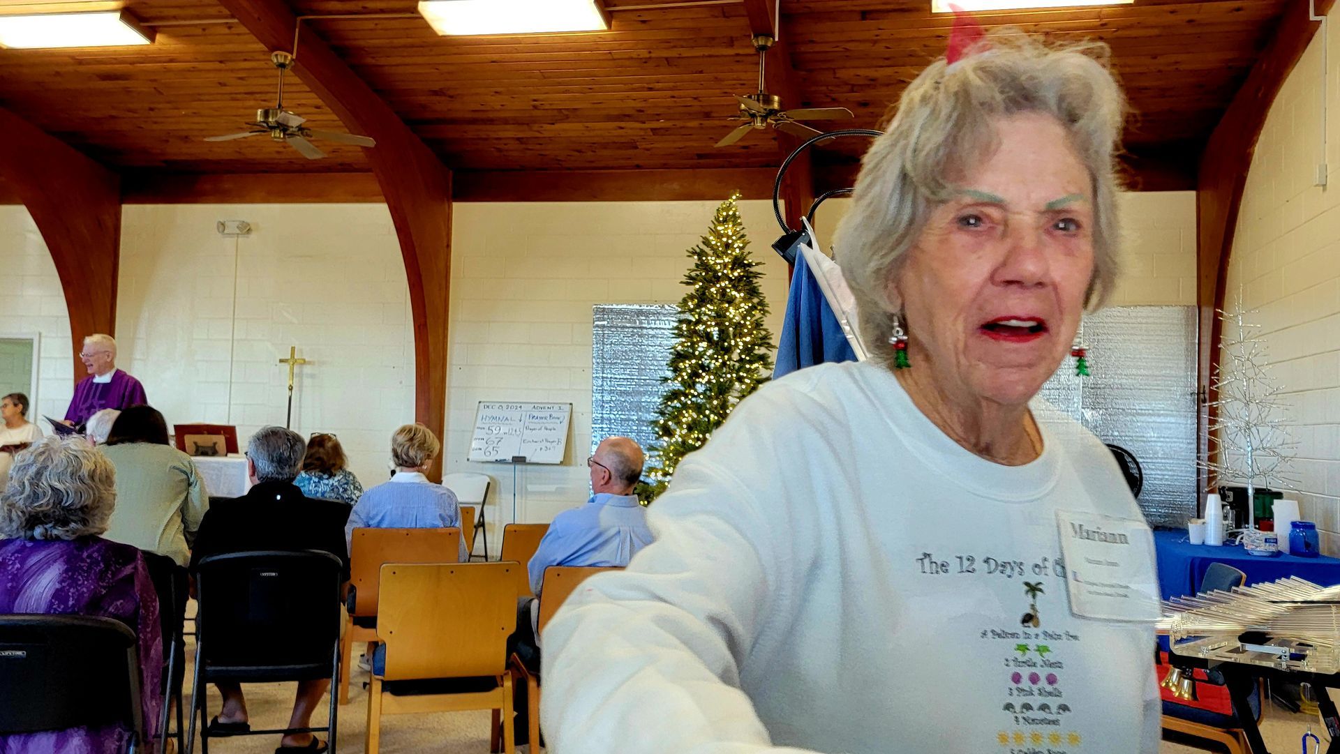 An elderly woman in a white sweatshirt looks at the camera, in a room with a church service.