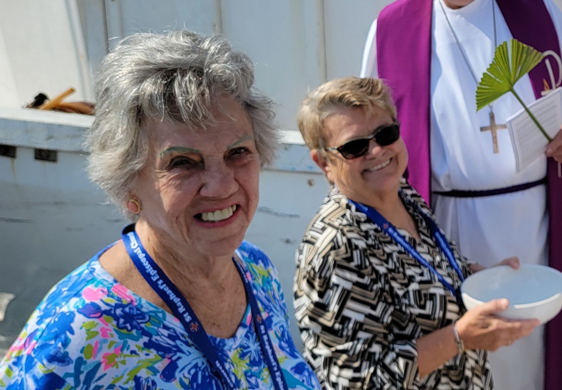 Two smiling women at an event, one in blue floral, the other in sunglasses, with a priest in purple.