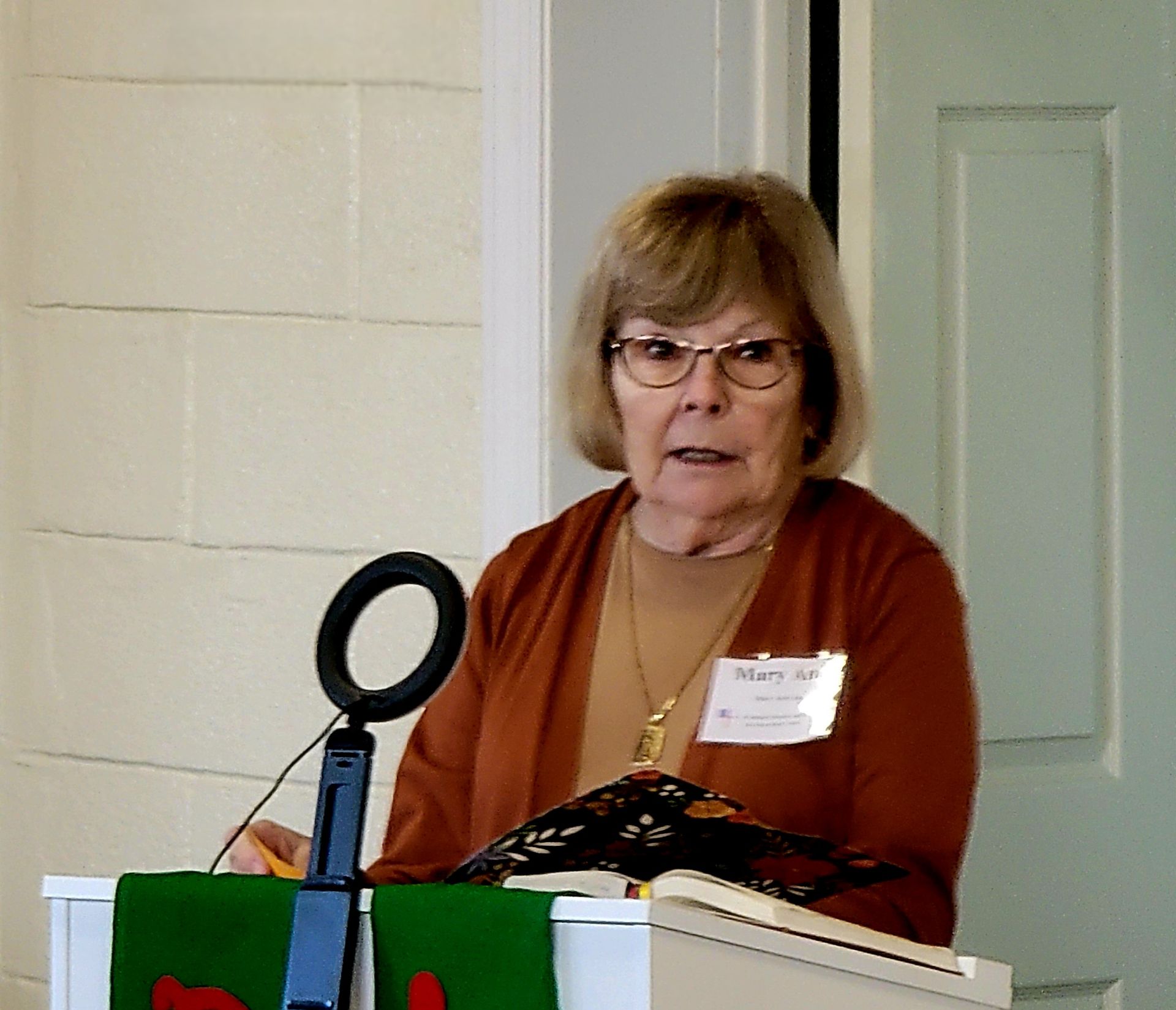 Woman speaking at a podium, wearing glasses and a brown cardigan. She has a microphone and name tag.