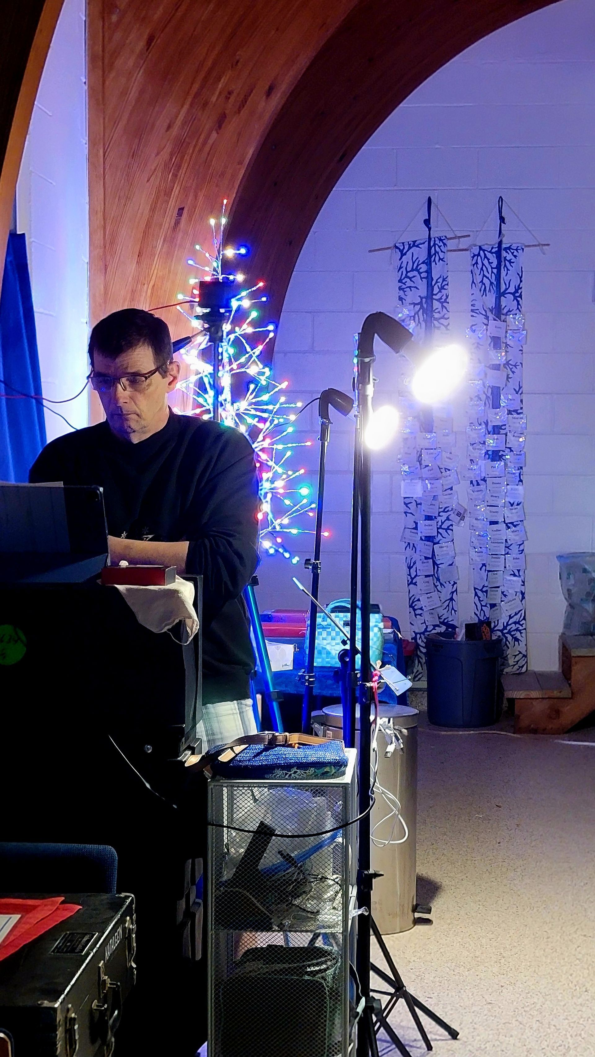 Man at a DJ setup with laptop, stage lights, and a Christmas tree in a room with blue and white lights.