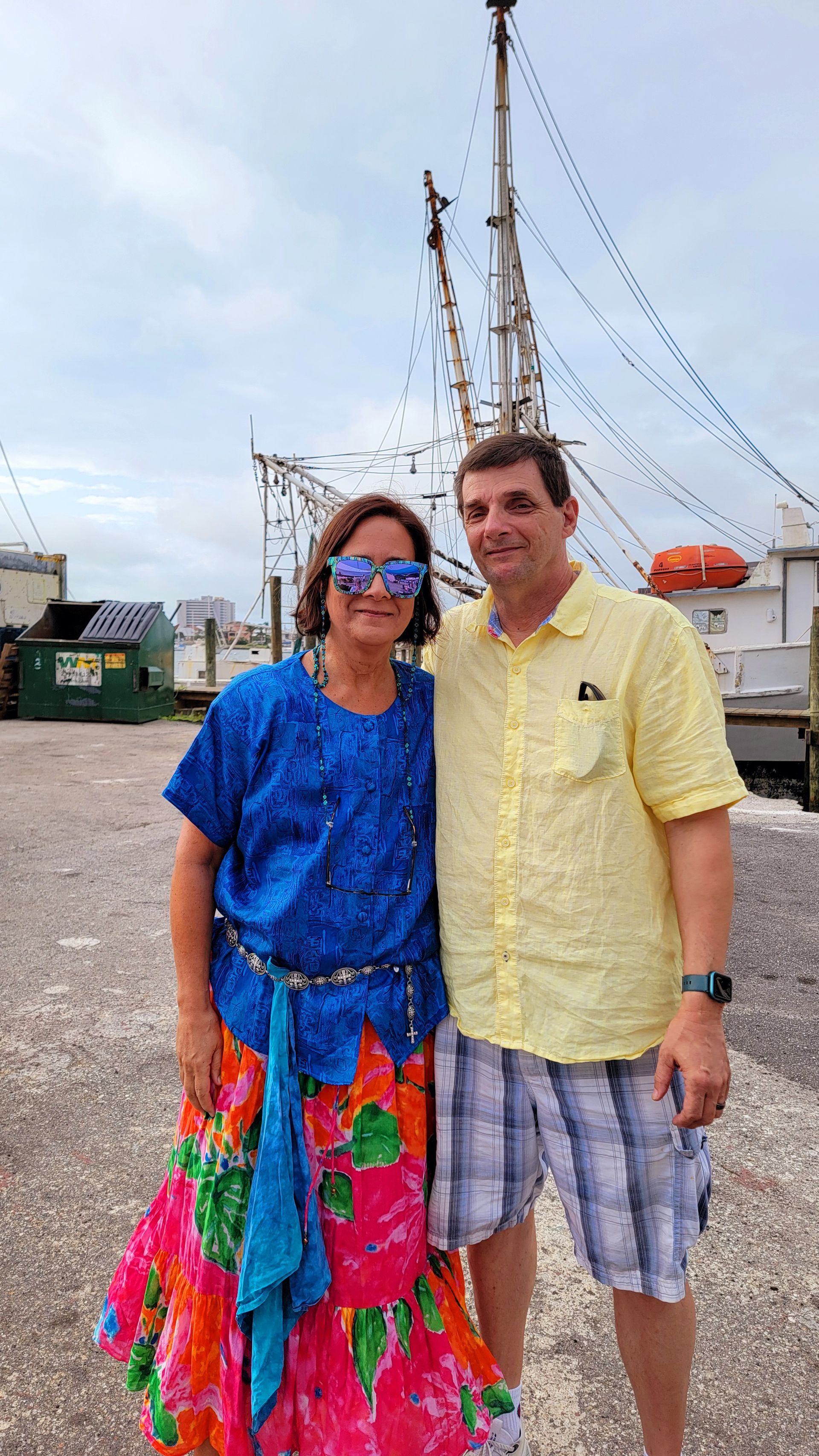 Woman in colorful outfit and man in yellow shirt and shorts pose by boats.
