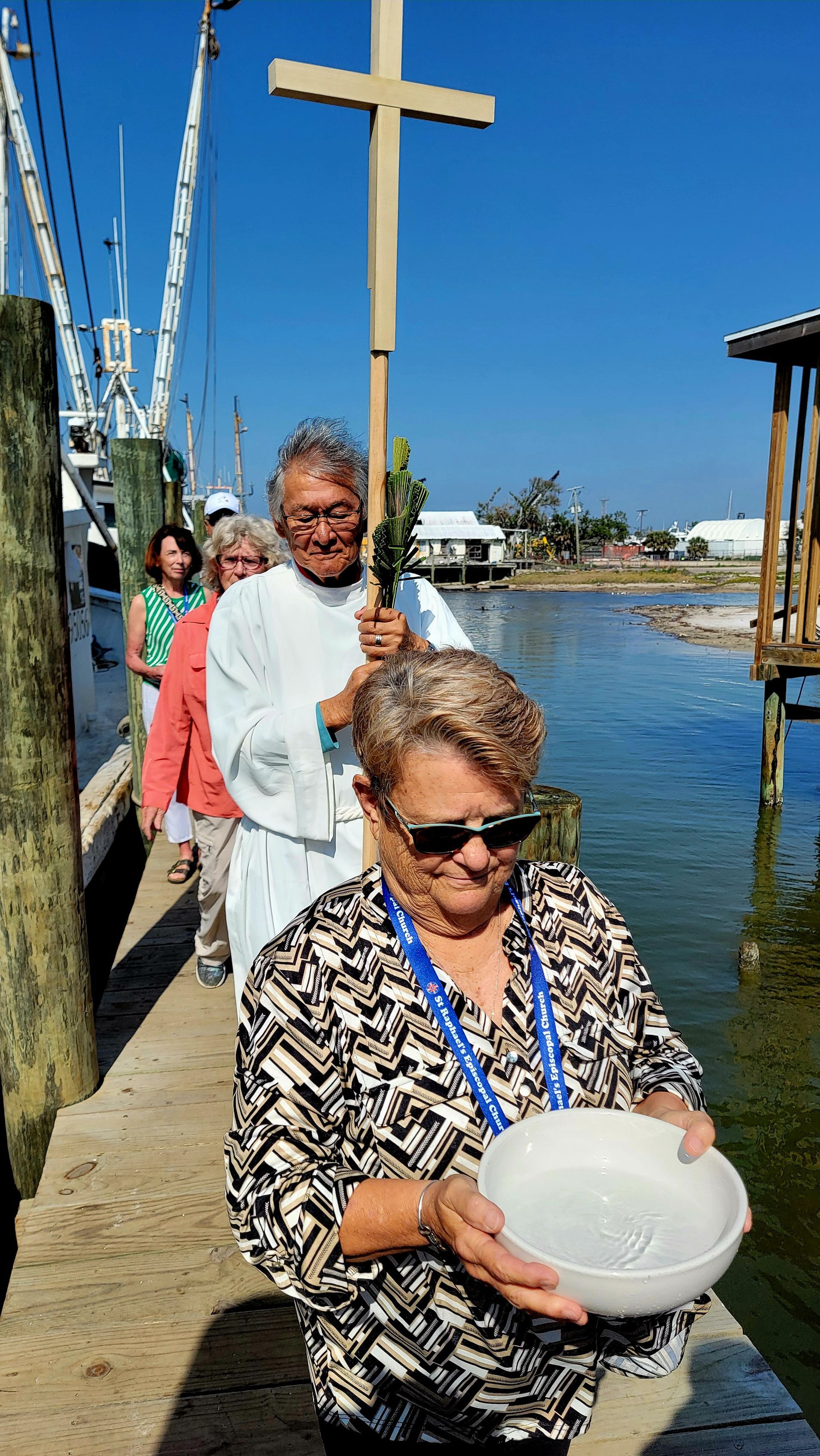 People in procession carrying a cross and bowl, walking along a dock near water.