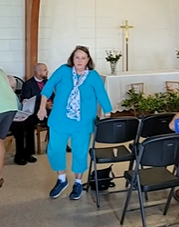Woman in blue outfit walks near chairs in a church setting, with a man in a suit and a cross in the background.
