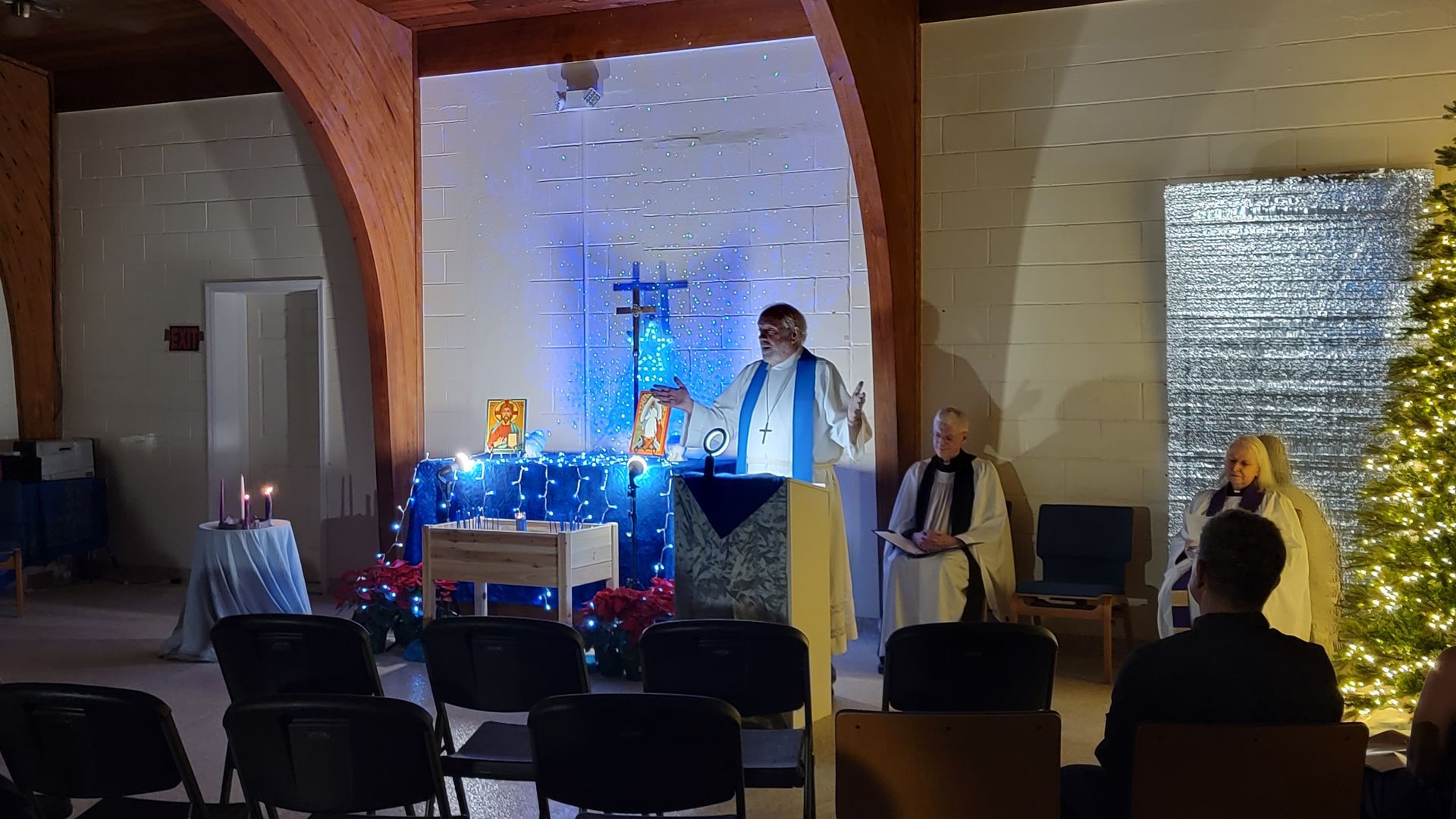A person in a blue stole speaking at a podium in a church, a cross and Christmas tree are visible.