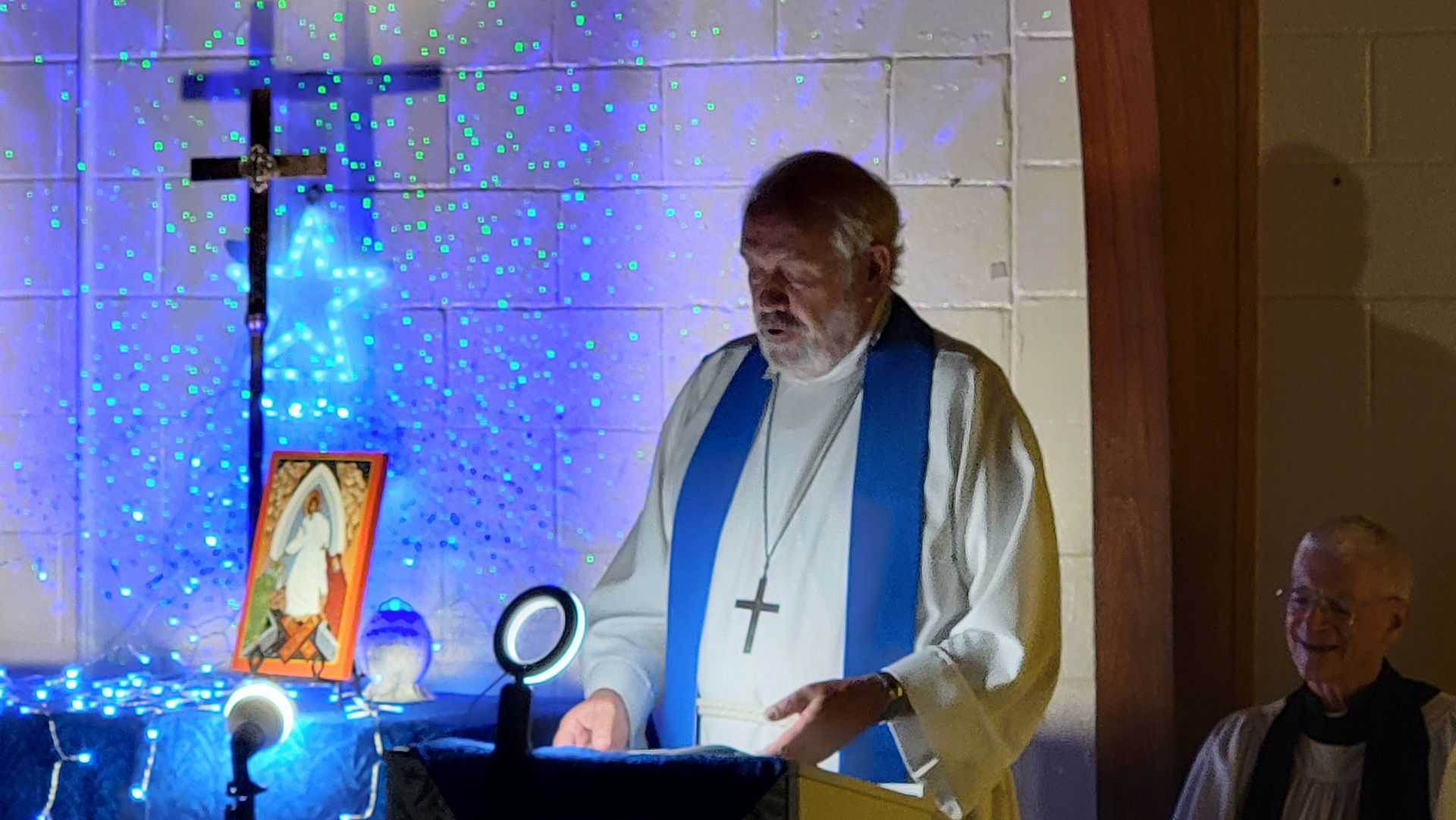 Man in robe with cross leading a service, altar decorated with lights, painting and a cross, another man in the background.