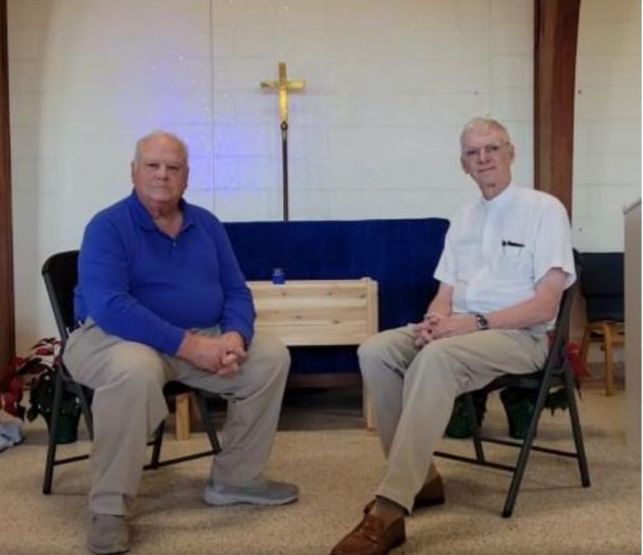 Two men seated in front of a cross. One wears blue, the other white. Church setting.