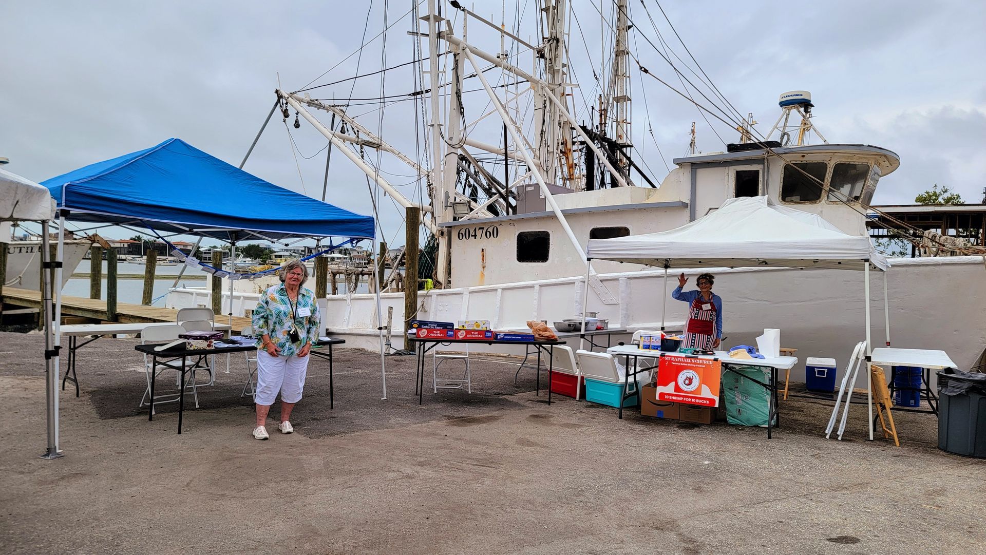 Woman and vendor at food stalls set up on a dock, behind a large white fishing boat under overcast skies.
