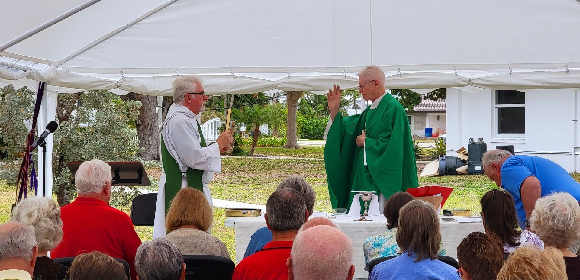 Two priests conducting a religious service under a white tent. Audience seated in front.
