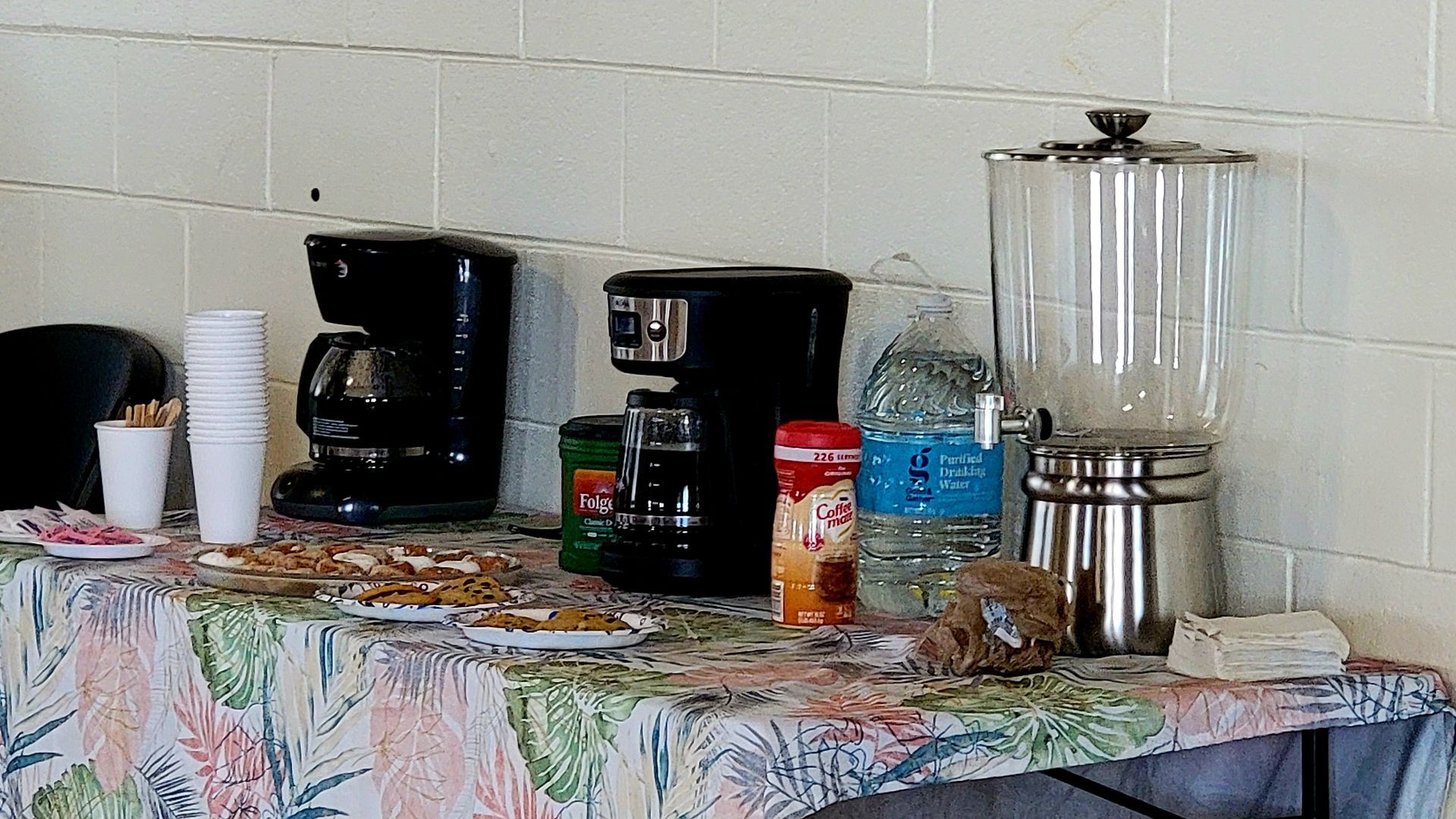 Coffee station with coffee makers, water, cups, and snacks on a floral tablecloth.