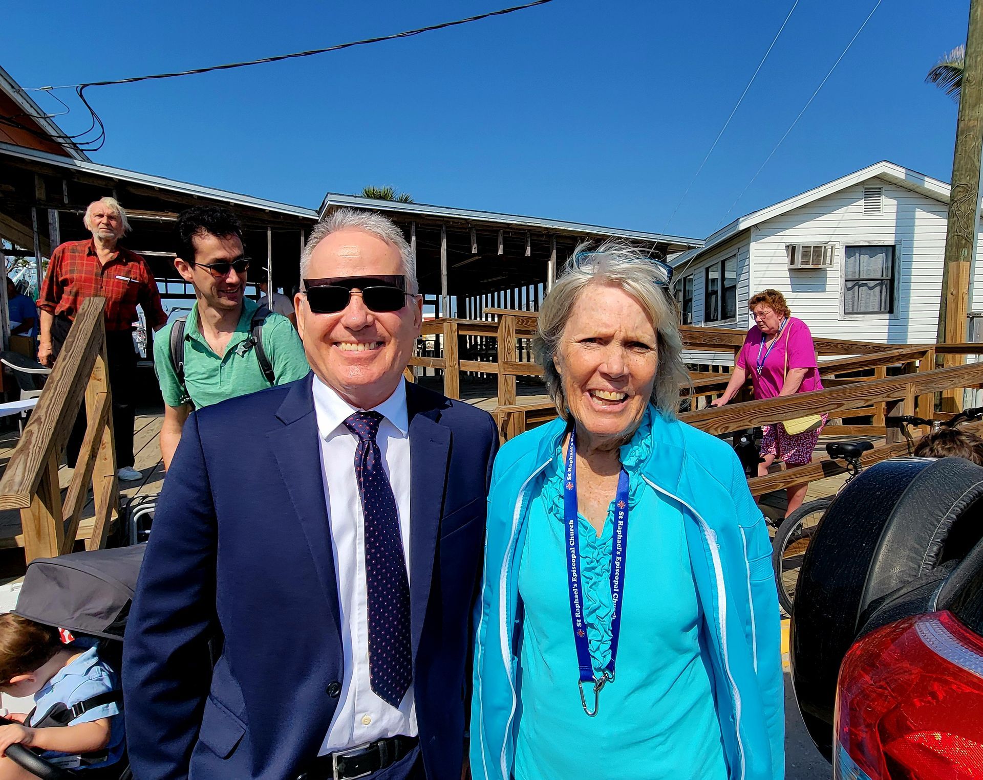 Man and woman smiling, posing outdoors near a building with other people visible. Blue sky.