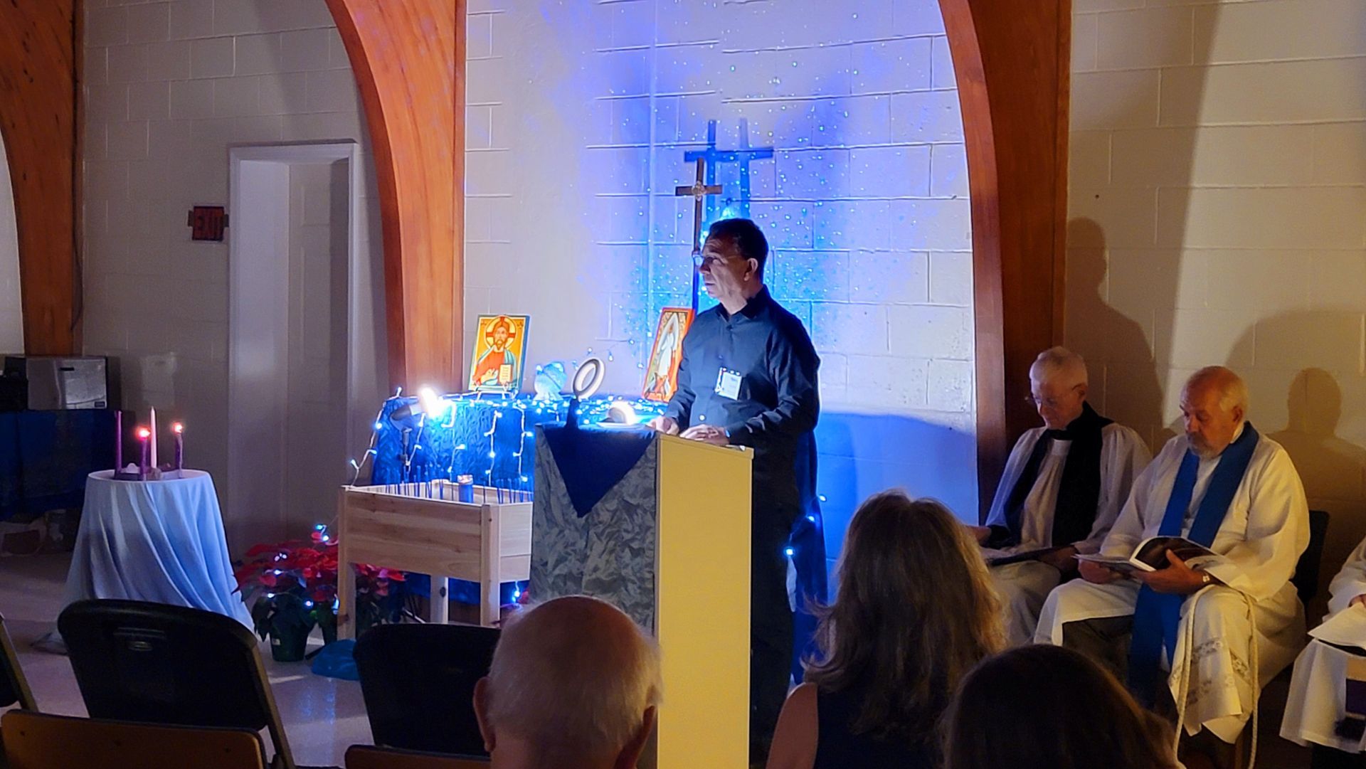 Man speaking at a podium in church with two clergy seated. Blue lights and cross in background.