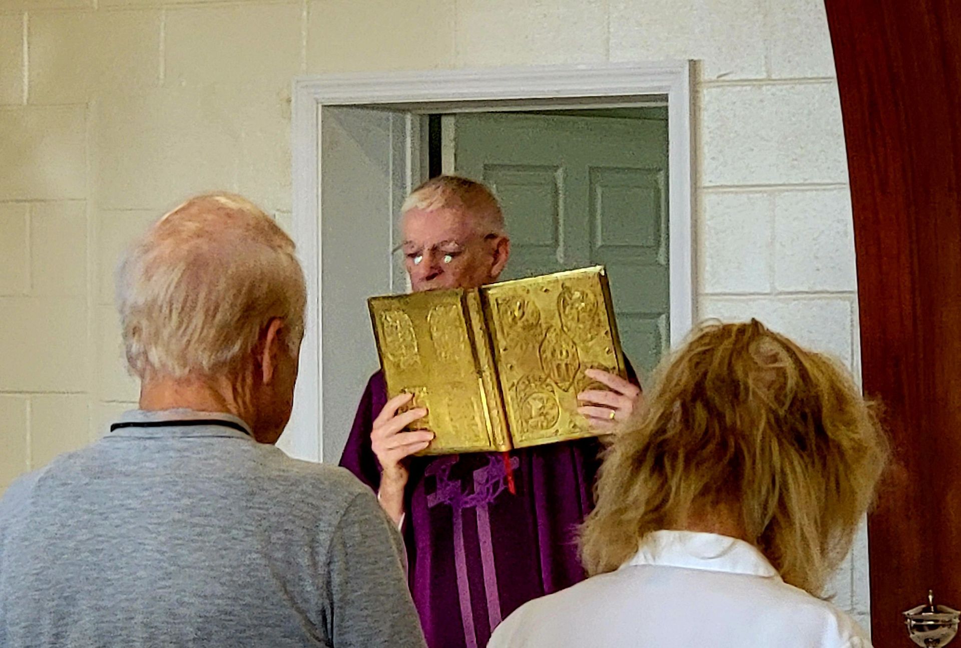 A priest in purple vestments holds a golden book open, facing two people in a doorway.