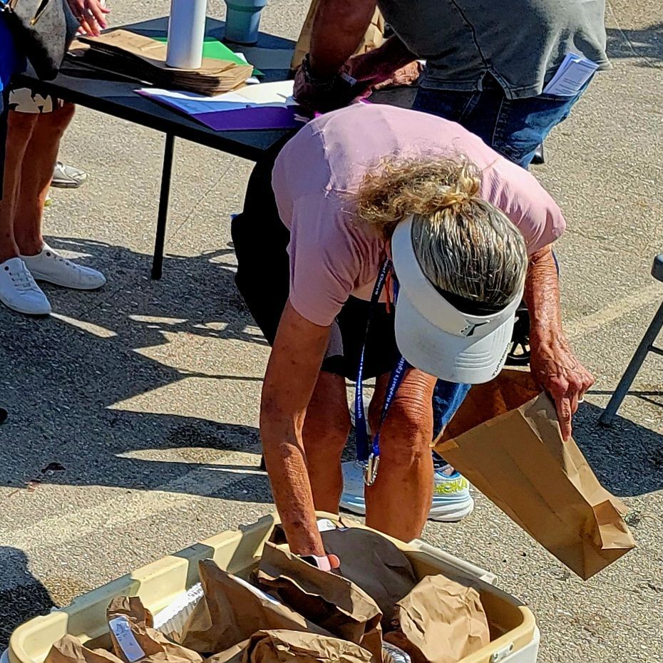 Woman reaching into a box of brown paper bags at an outdoor event. Others are nearby.