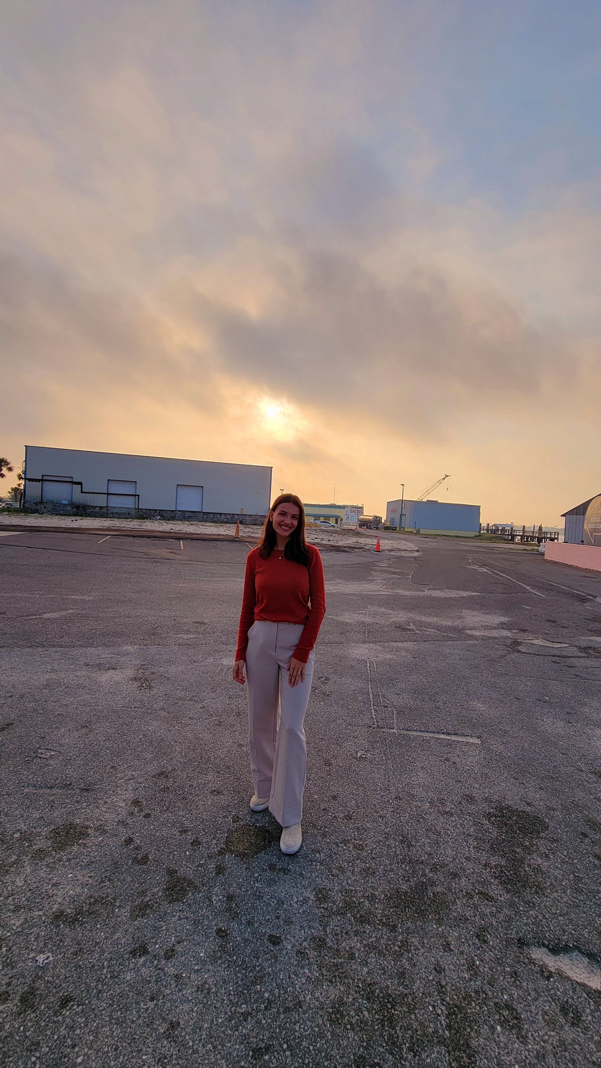 Woman standing on a pebble beach at sunset. Wearing red sweater and white pants, buildings in background.
