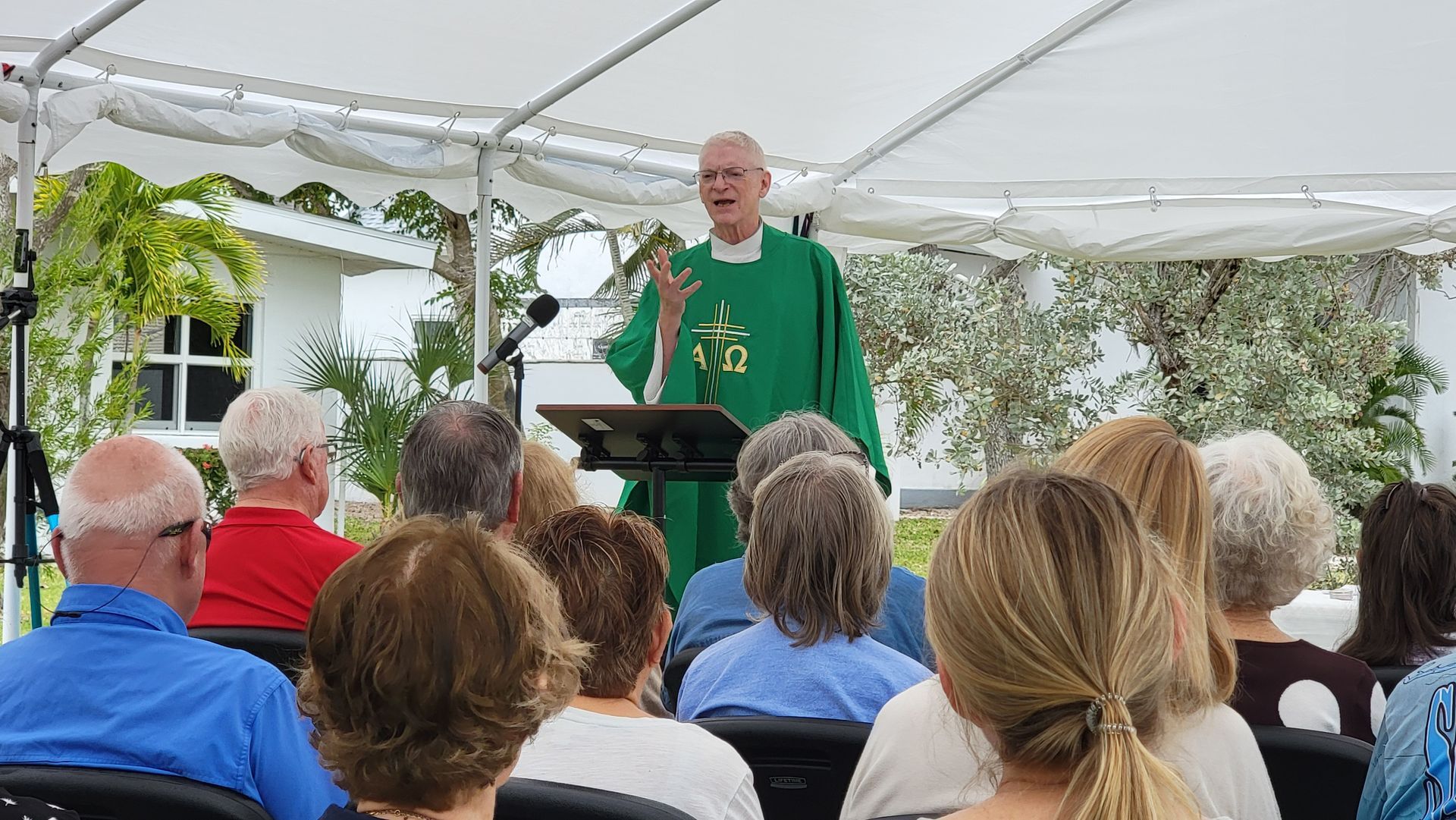 Priest in green vestments speaking outdoors to seated congregation under tent.