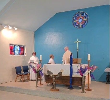 Altar in a church with a priest, a server, and a lector during a service. Blue wall, stained glass.