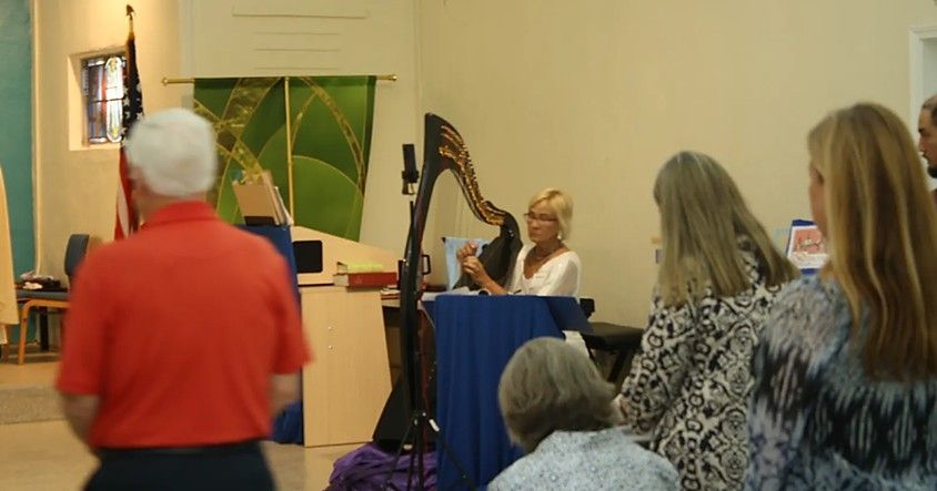 Woman playing harp in front of a small audience in a church-like setting.