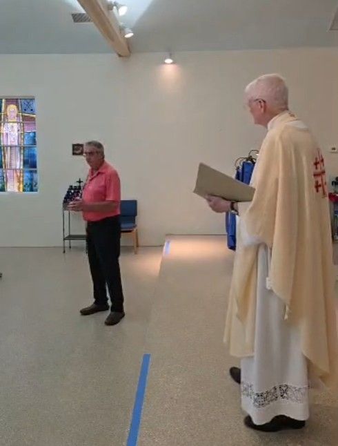 A man in a pink shirt speaks. A robed person holds a book. In a room with a blue line and a stained-glass window.