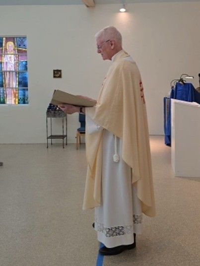 Priest in white vestments reads from a book, standing in a church with a stained glass window.