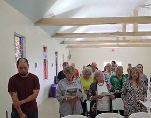 People in a room, some holding papers, heads bowed. Beige walls, stained glass windows, and wood beams.