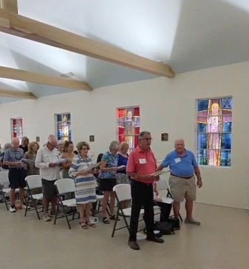 People in a room, holding papers, possibly singing. Stained glass windows visible.