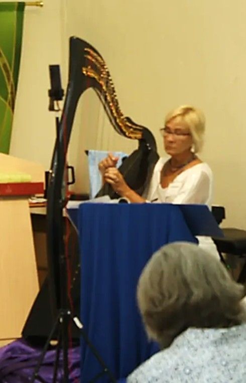 Woman playing a black harp. She is adjusting strings, wearing glasses, and a white top, seated behind a blue table.