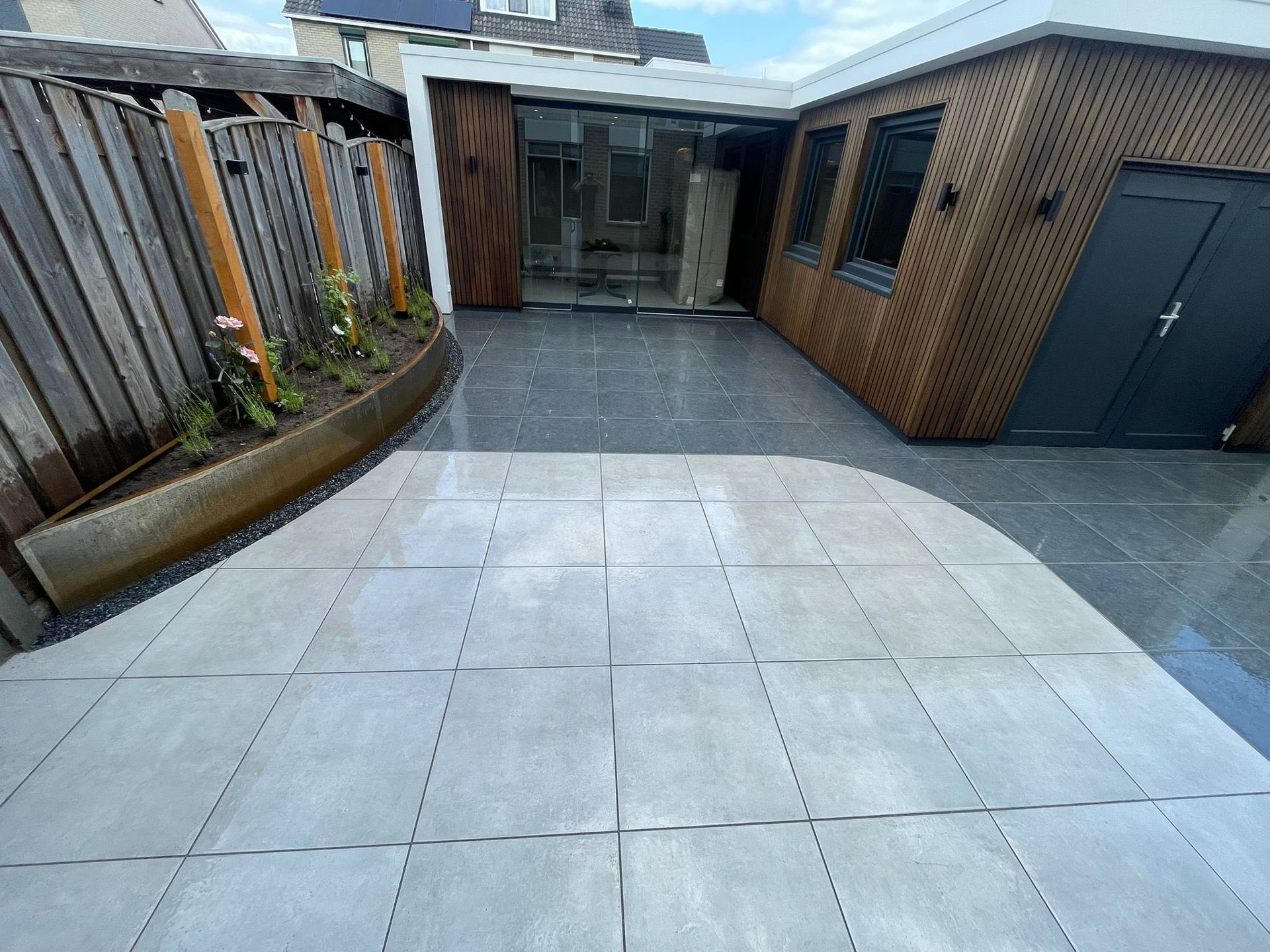 Een terras in de achtertuin met lichtgrijze vierkante tegels, een houten schutting met plantenbakken en een moderne aanbouw met glas.
