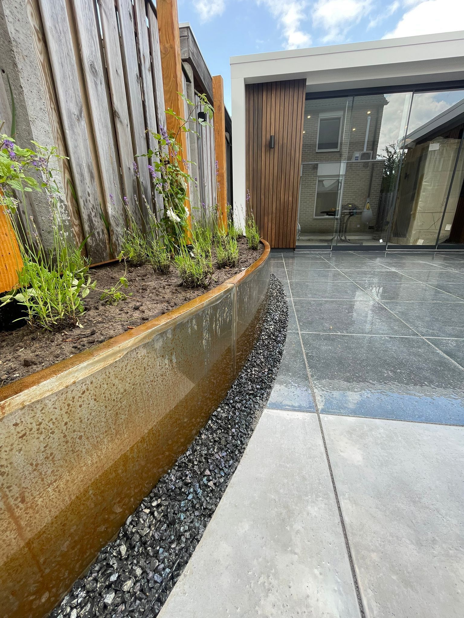 Een gebogen, verroest metalen plantenbed gevuld met kleine plantjes, naast een zwarte grindrand en een lichtgrijs terras.