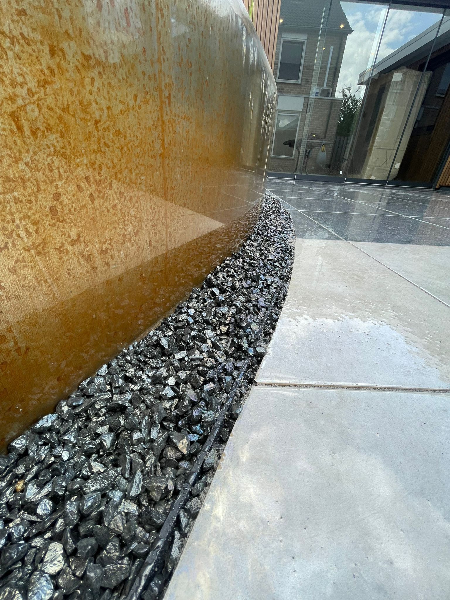 Een metalen muur met een roestige afwerking staat naast een strook donkere, gebroken steen en lichtgekleurde betonnen bestrating.