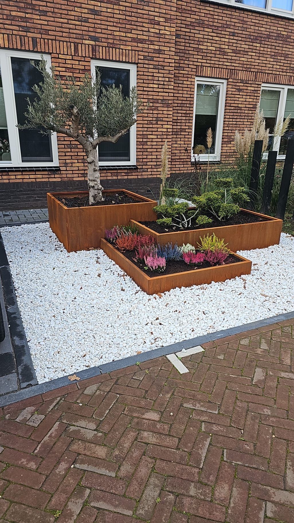 Een plantenbak van cortenstaal met meerdere niveaus, gevuld met een kleine boom en bloemen, geplaatst in een tuin met witte stenen en bakstenen bestrating.