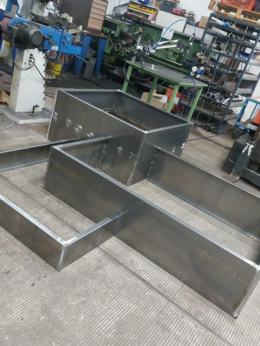 Drie gelaagde plantenbakken van cortenstaal met een olijfboom en kleine bloemen, geplaatst in een wit kiezelbed naast een bakstenen gebouw.