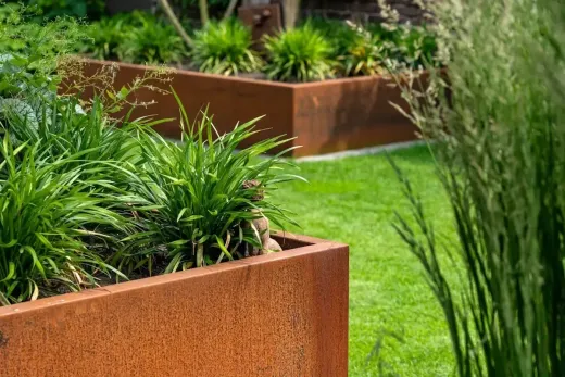 Verhoogde plantenbakken van cortenstaal, gevuld met groene planten, staan ​​in een achtertuin met gazon.