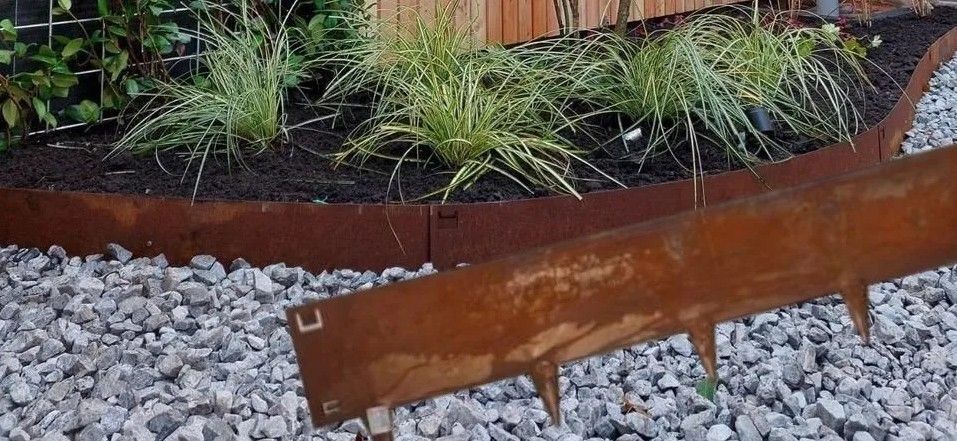 Een roestige metalen afbakening scheidt een bloembed met siergrassen en mulch van een pad van grijs grind.
