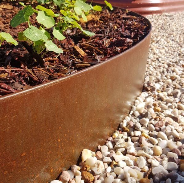 Een close-up van een verroeste metalen tuinrand die houtsnippers en kleine groene plantjes scheidt van lichtgekleurd grind.