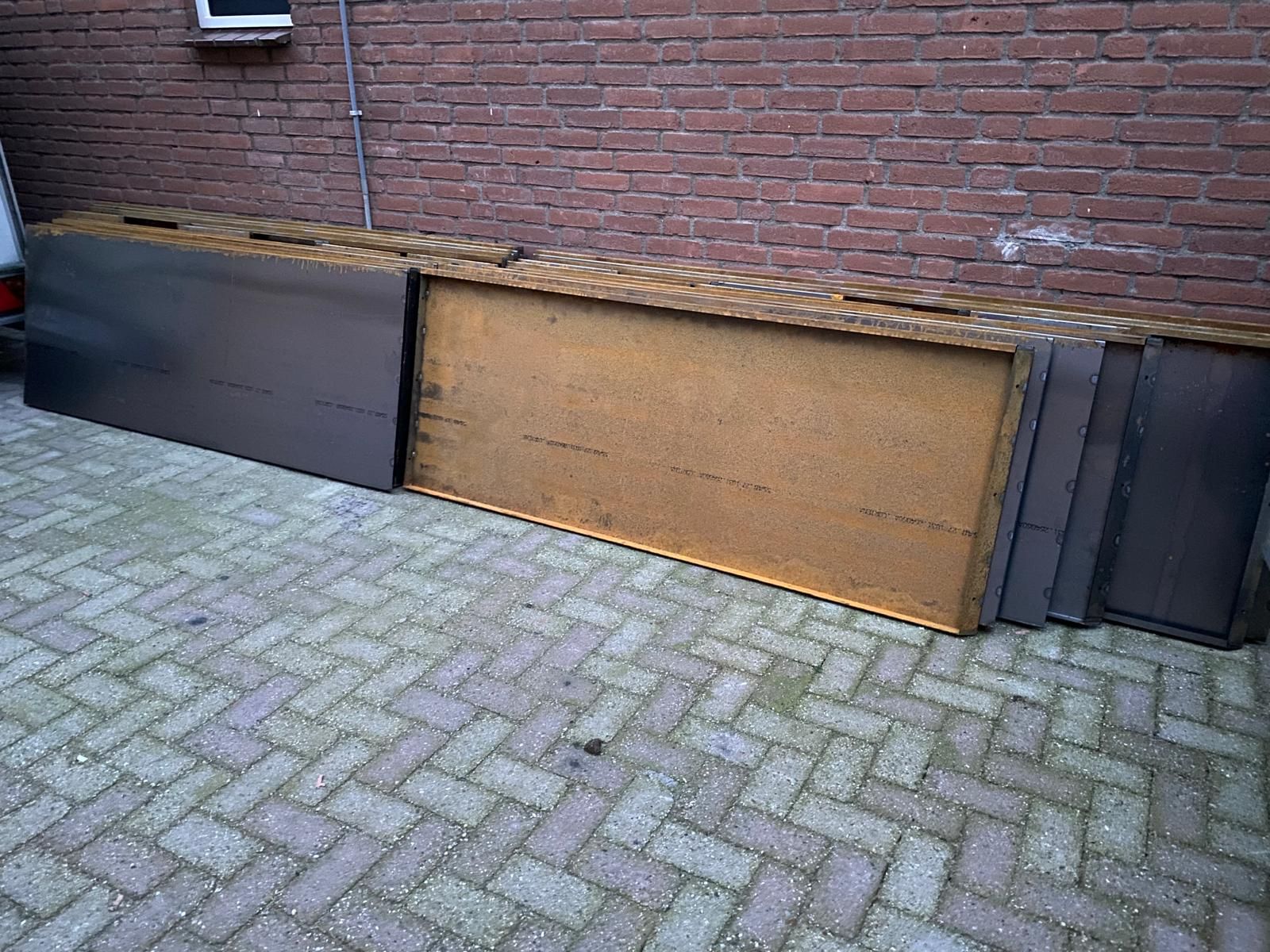 Verschillende rechthoekige houten panelen met een donkerbruine afwerking staan ​​tegen een bakstenen muur op een betegeld terras.