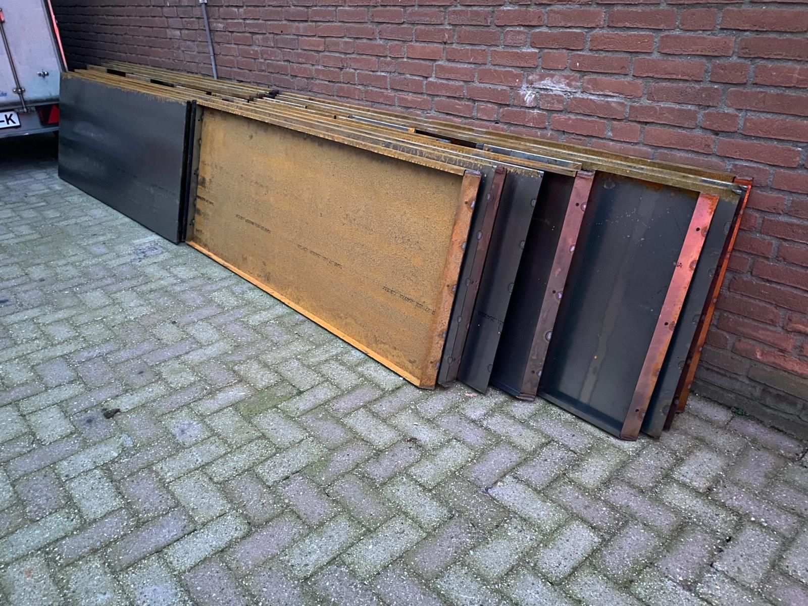Een rij van meerdere rechthoekige bekistingspanelen die tegen een bakstenen muur op een bestrate ondergrond leunen.