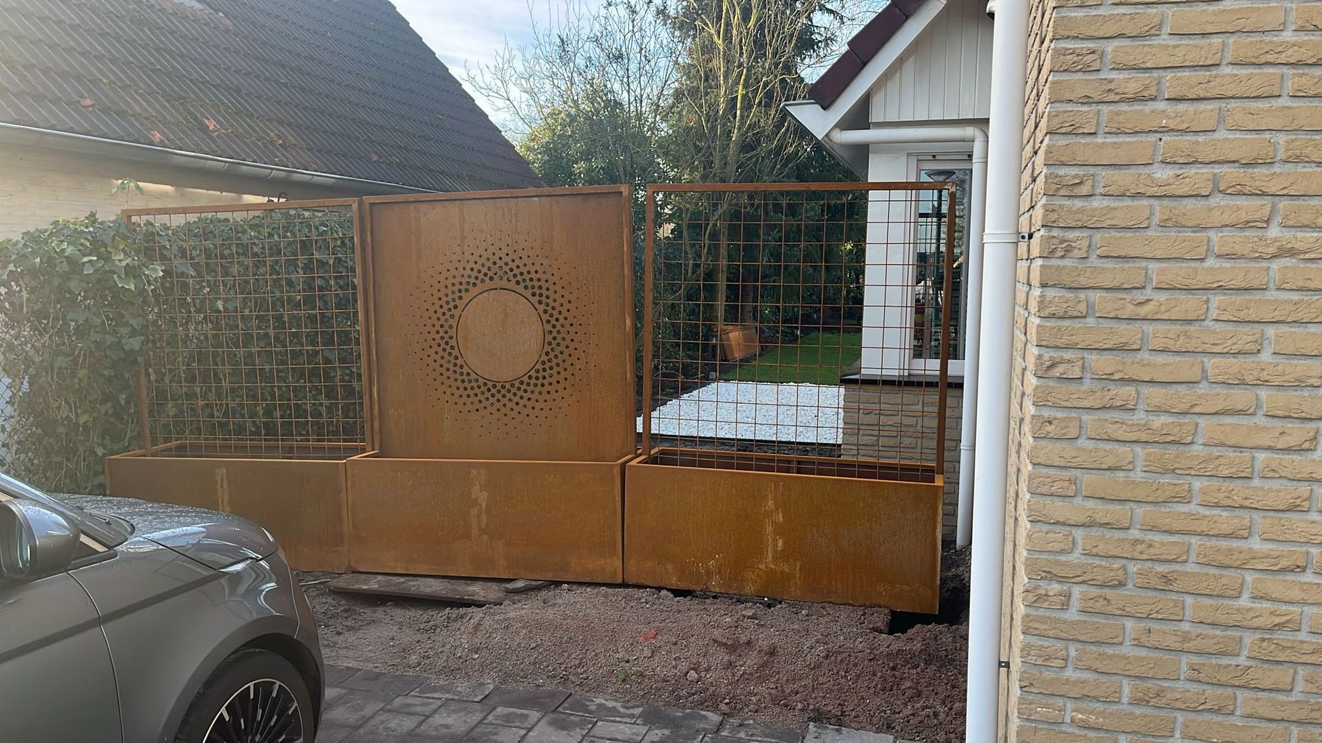 Decoratieve, roestkleurige metalen privacyschermen en plantenbakken zijn geplaatst in de oprit naast een woonhuis.