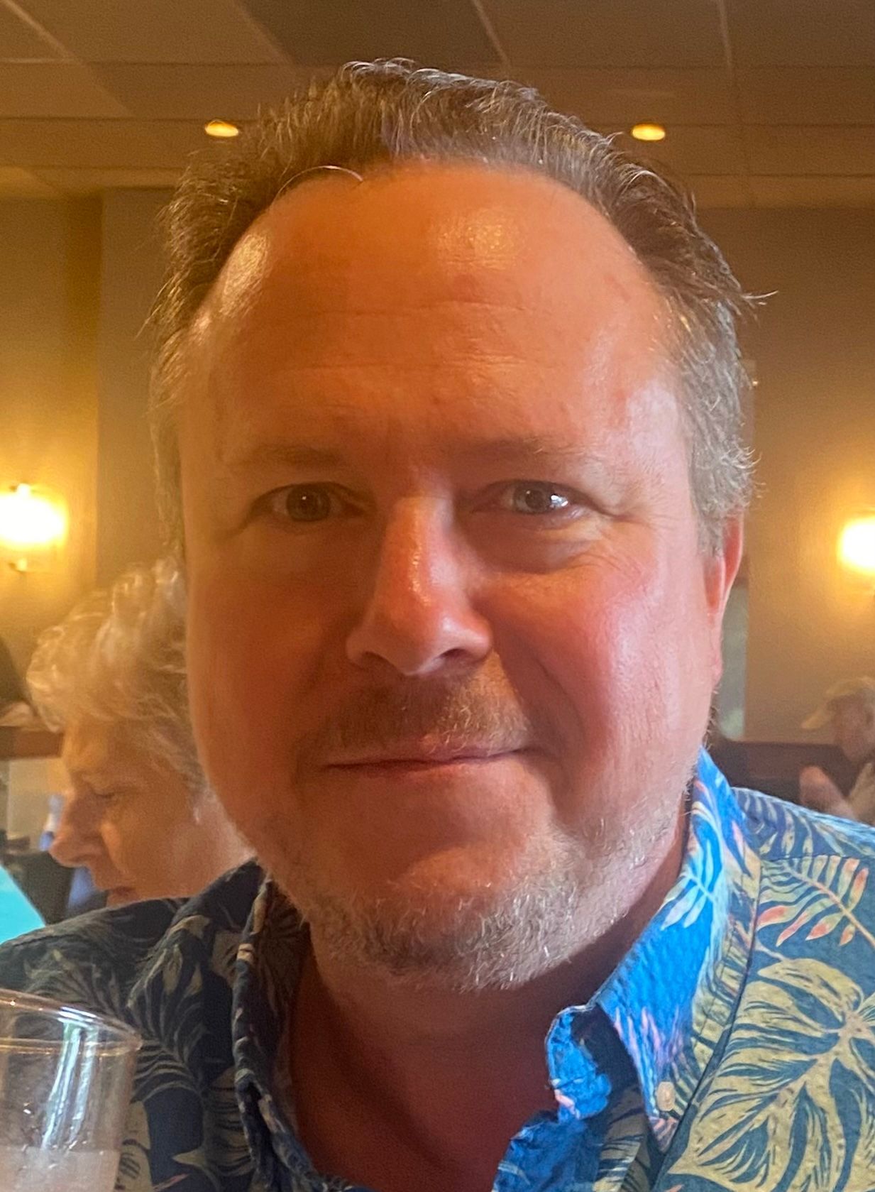 Man smiling in a blue patterned shirt in a warmly lit indoor setting