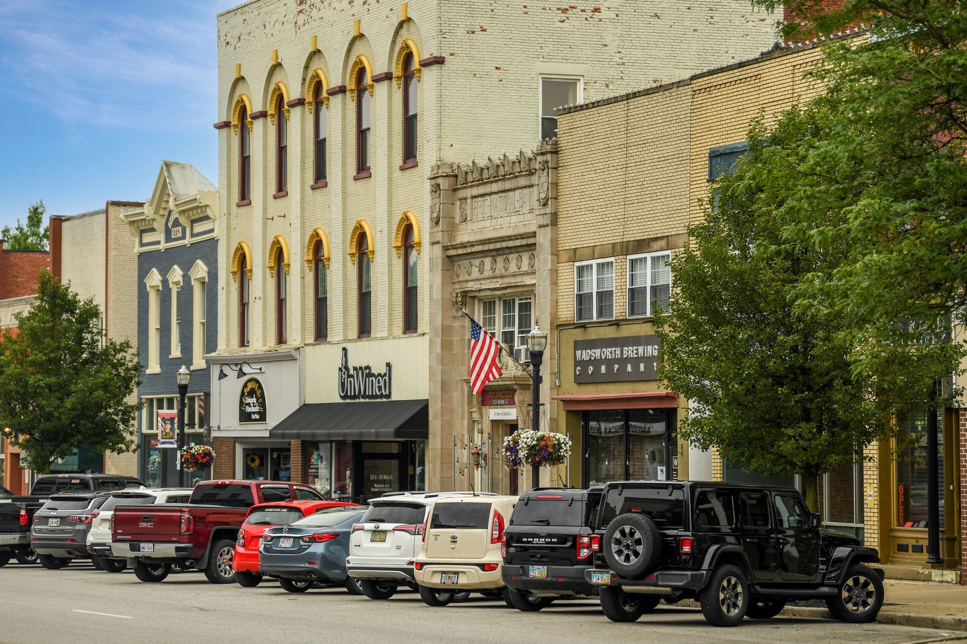 Downtown street with historic storefronts, parked cars, and trees in front of ornate buildings
