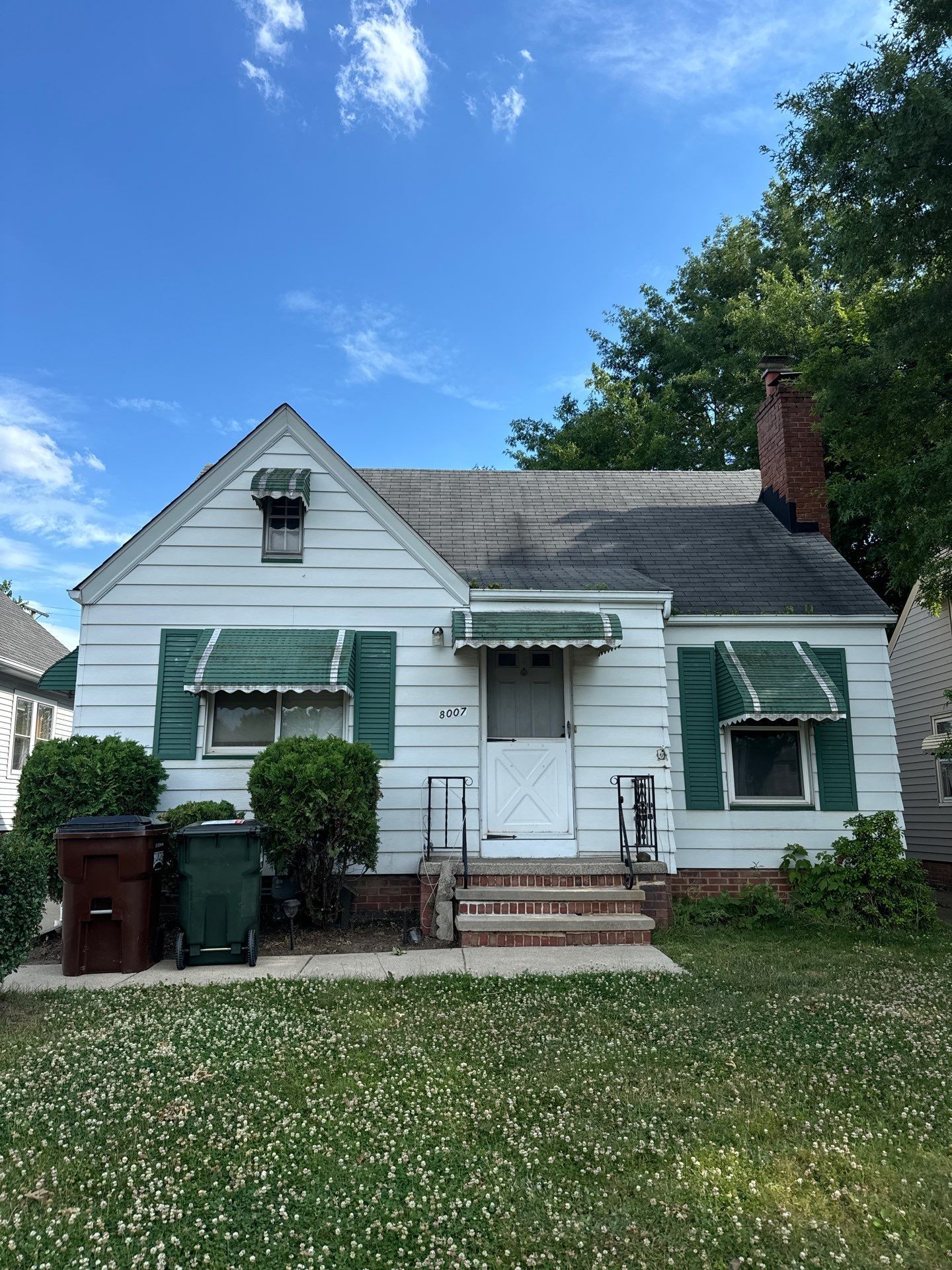 Small white house with green awnings, brick steps, and a front yard under a blue sky