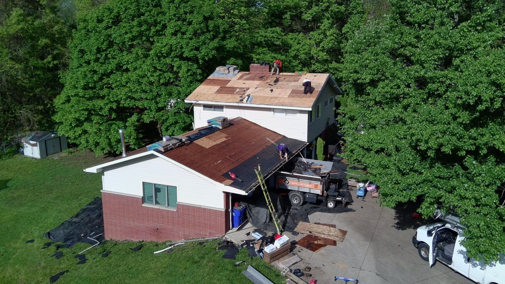 Aerial view of a damaged house with a collapsed roof and debris beside dense green trees.