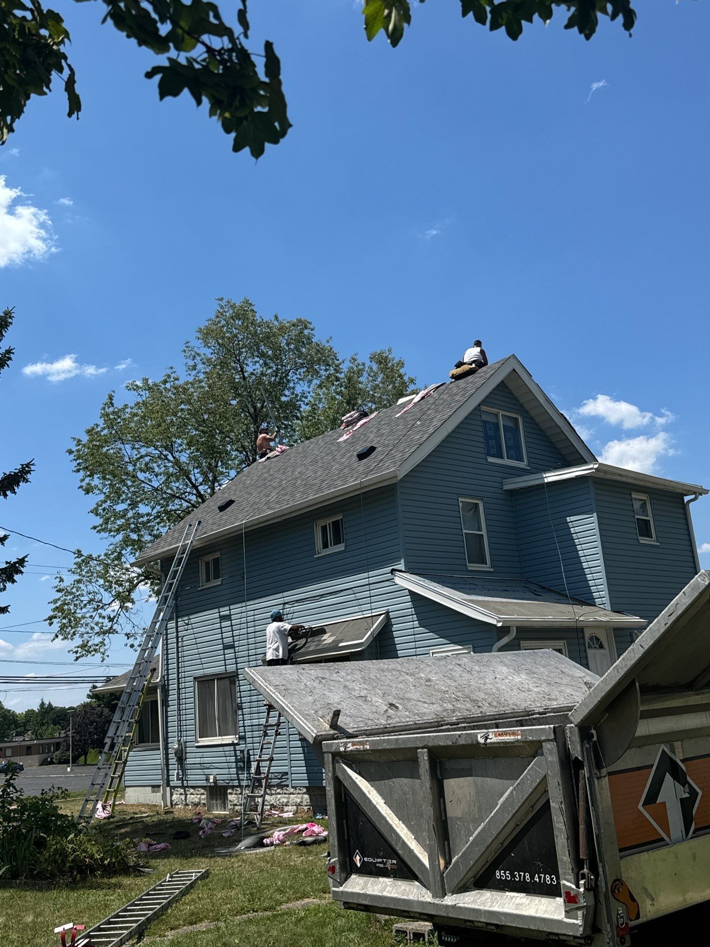 Blue two-story house with gray roof, surrounded by trees under a sunny sky, with a small shed in front.