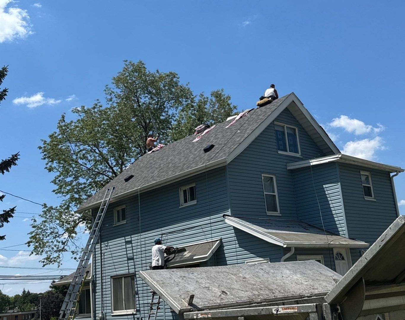 Blue house with damaged roof shingles and debris after storm under a clear sky