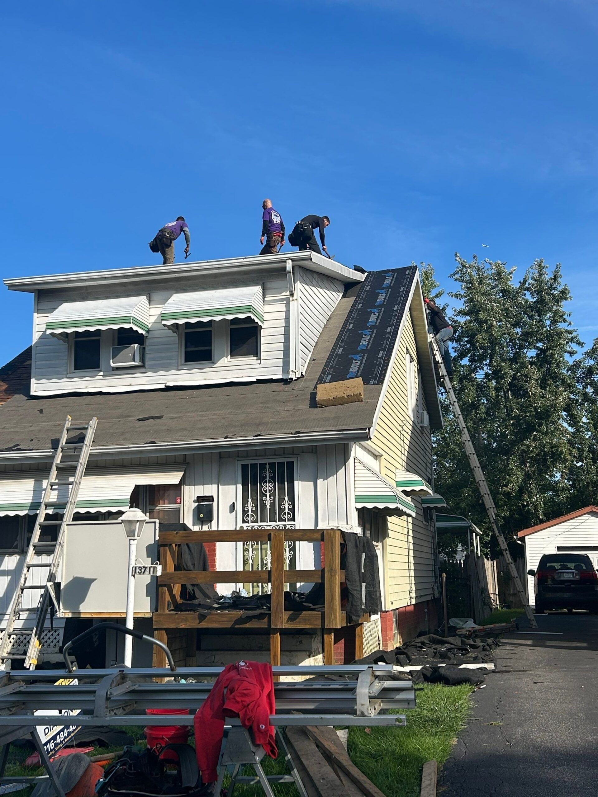 Workers on a roof repairing storm damage on a white two-story house with a damaged side wall.