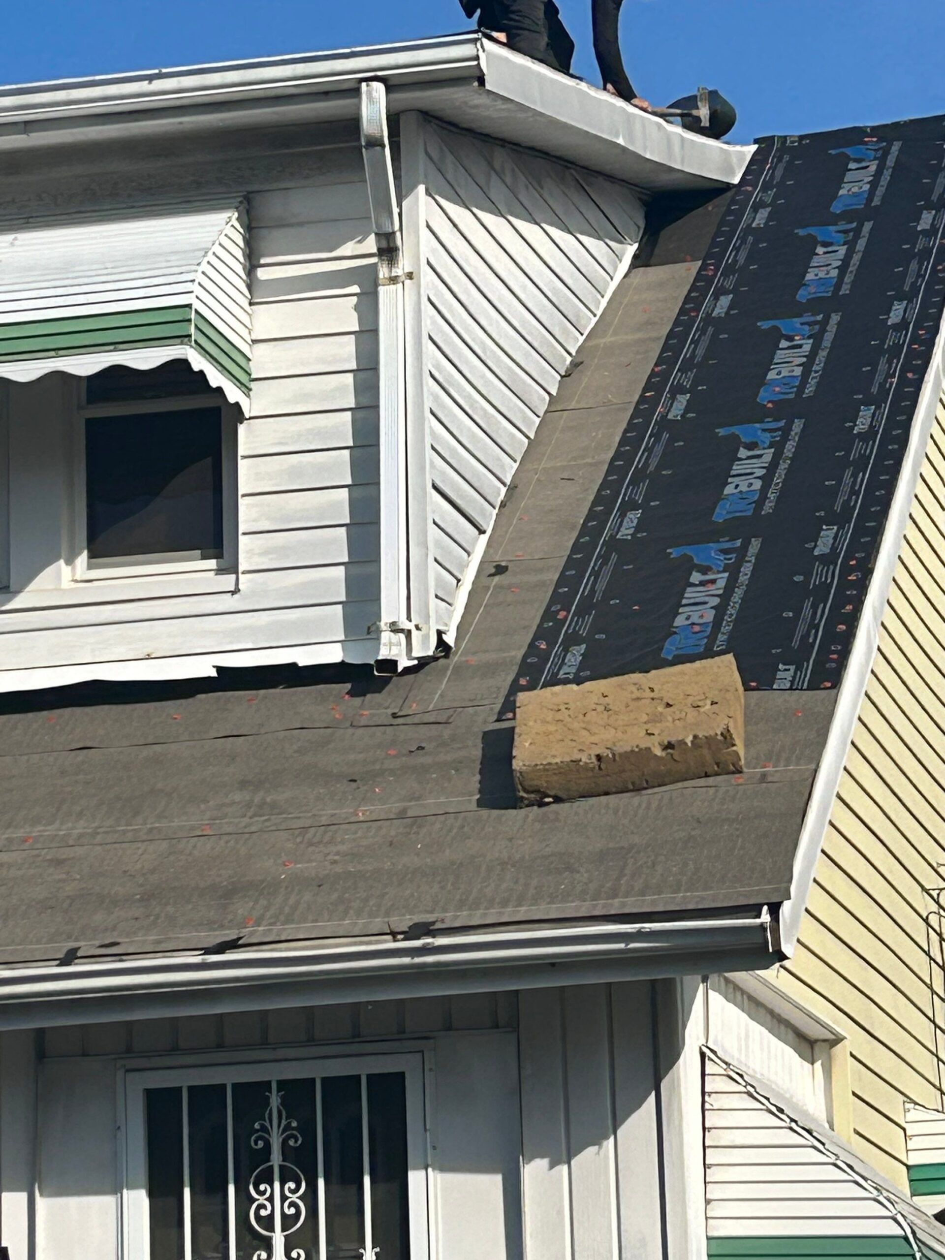 Roof shingles being installed on a house, with black underlayment and a yellow siding wall visible.