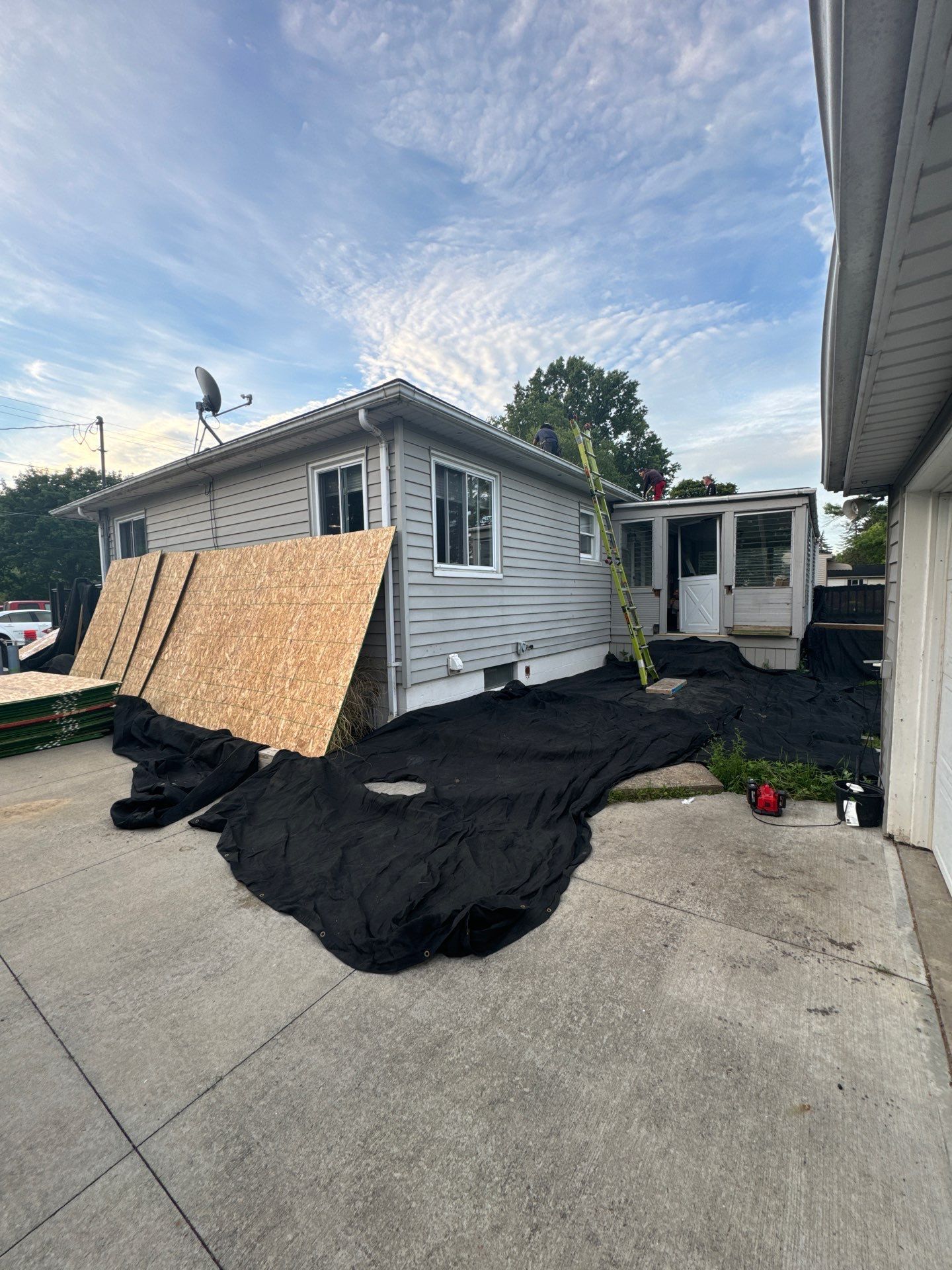 House under repair with plywood covering windows and black tarp on driveway under a cloudy sky