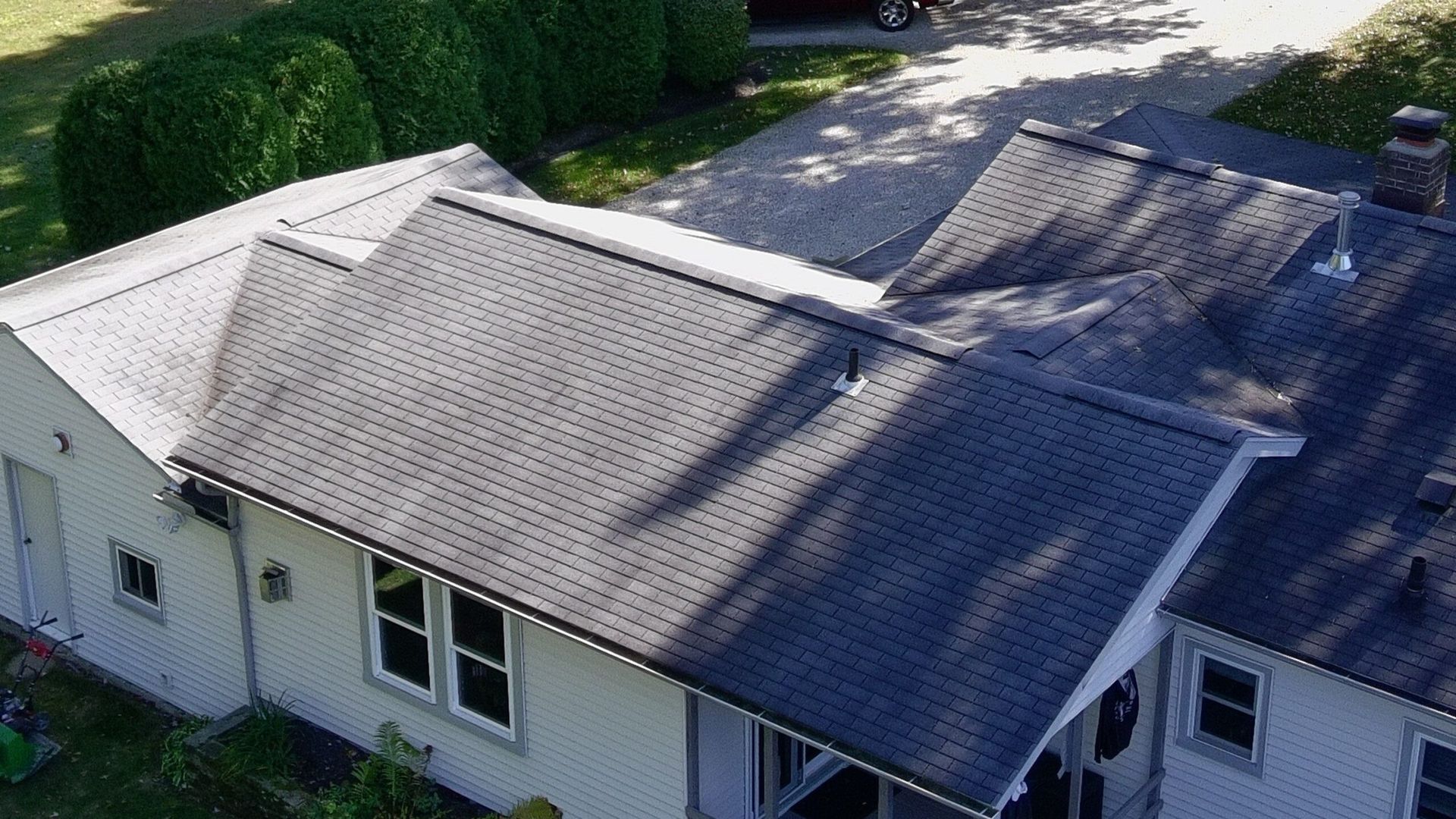 Aerial view of a house roof with gray shingles and white siding, surrounded by greenery.