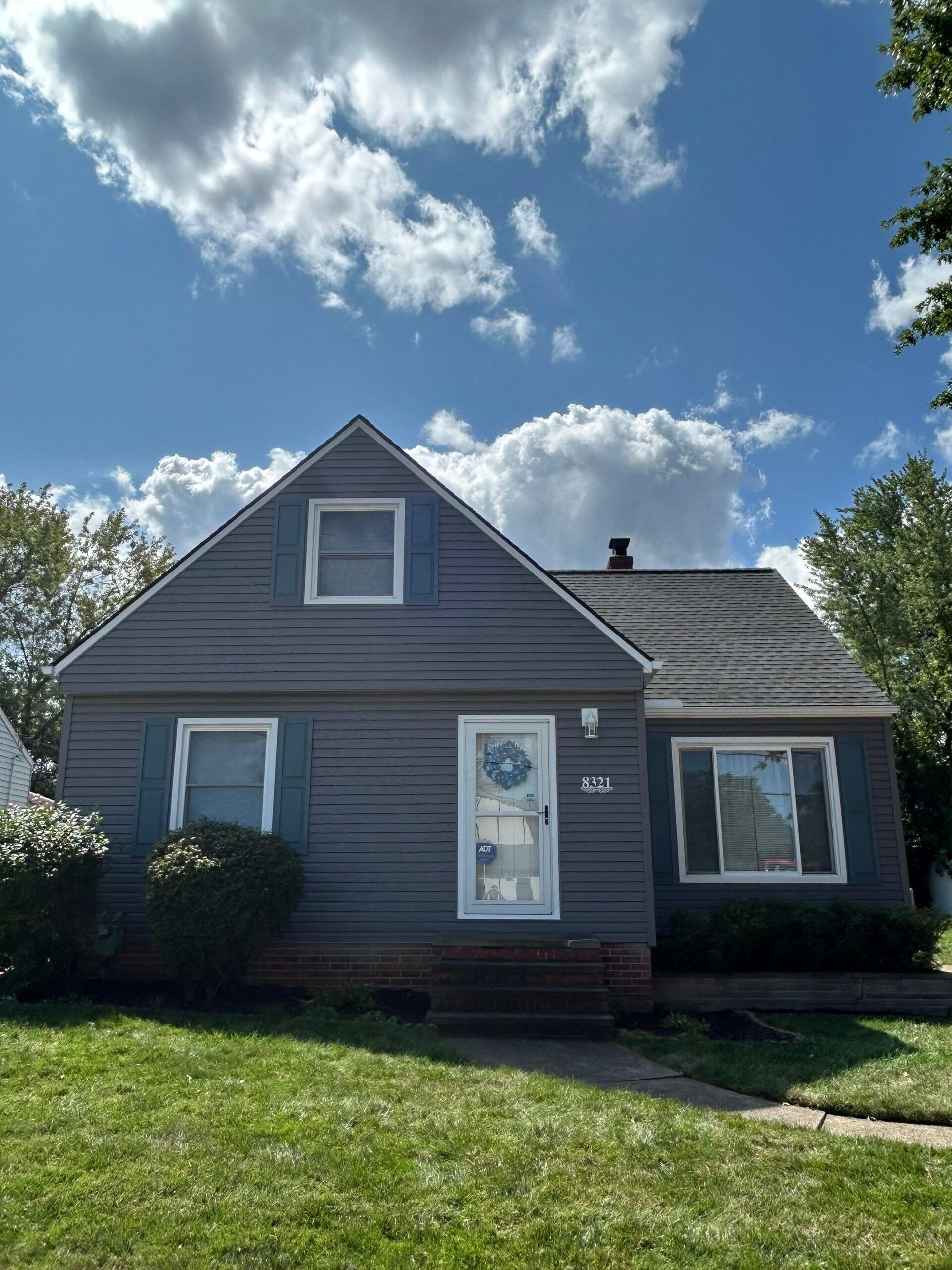 Gray house with white trim and a front door, viewed from the lawn under a blue sky with clouds