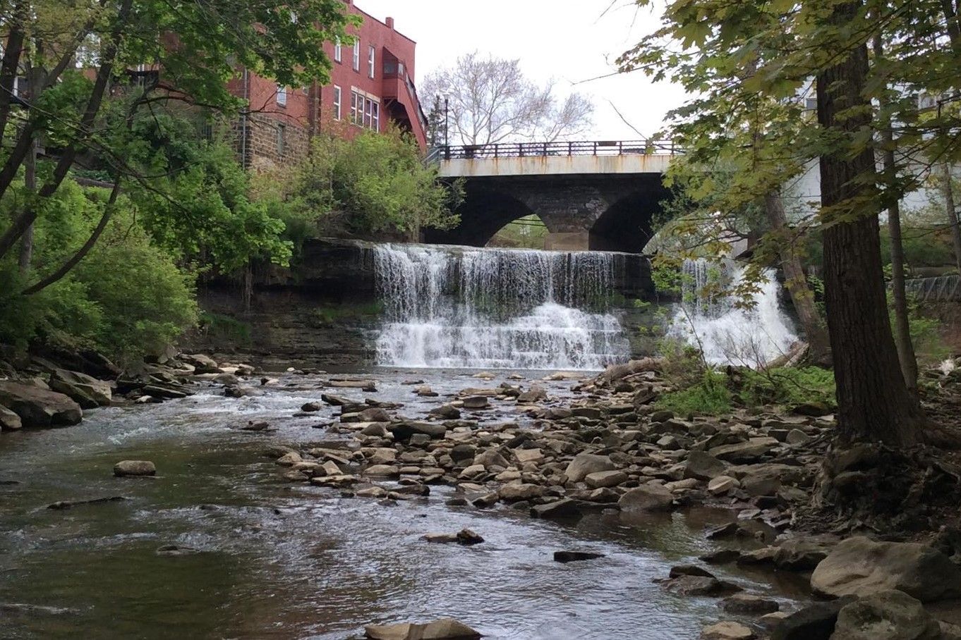 Waterfall in a wooded park beneath a bridge, with rocks and a red brick building nearby