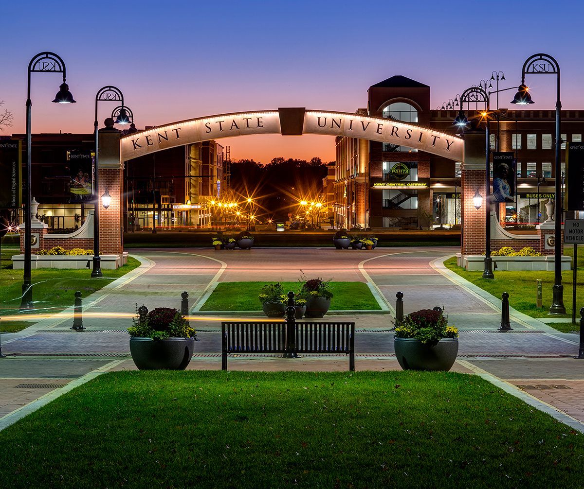 Illuminated campus entrance at dusk with arched gateway, lamp posts, benches, and landscaped lawn.